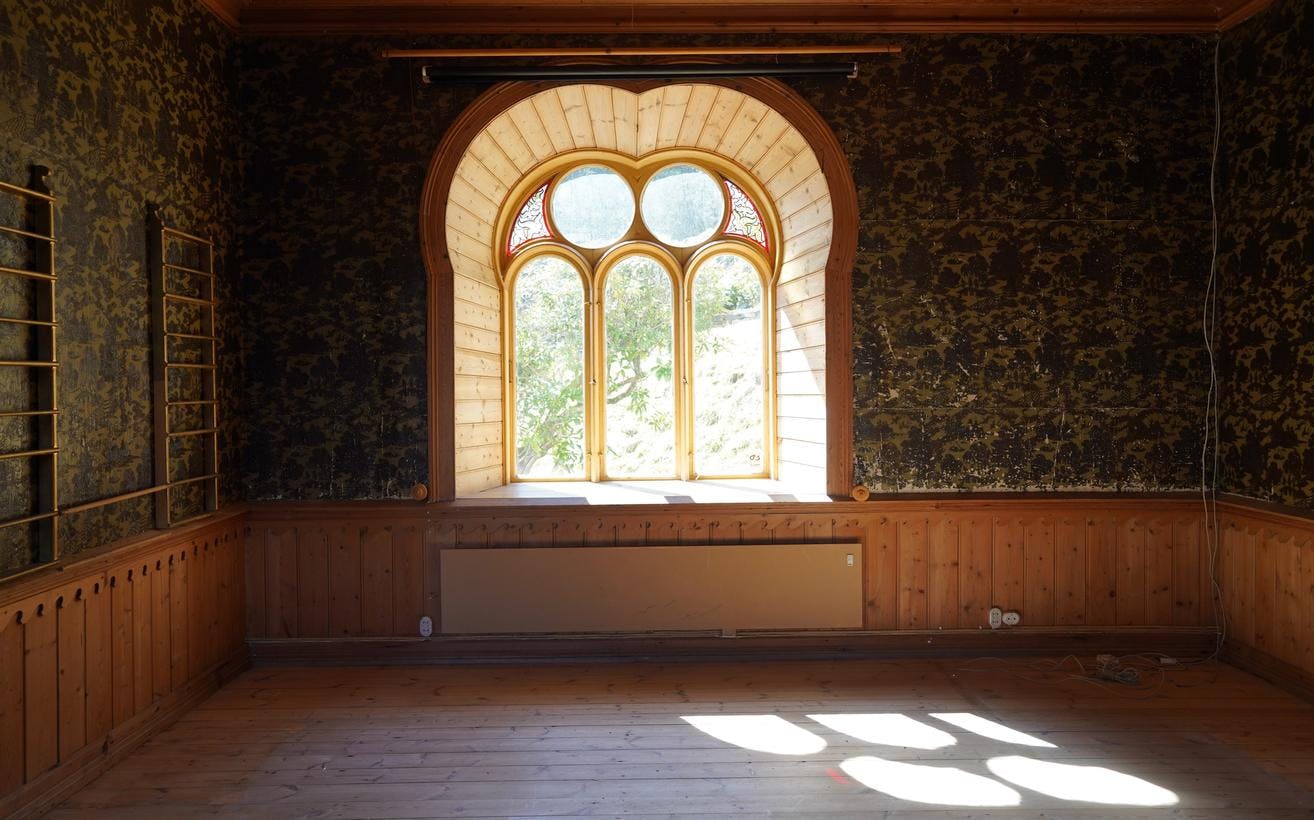 Et tomt rom i villaen på Lysøen, med et stort vindu midt på veggen, hvor lyset faller inn. Rikt ornamentert tapet på veggene.