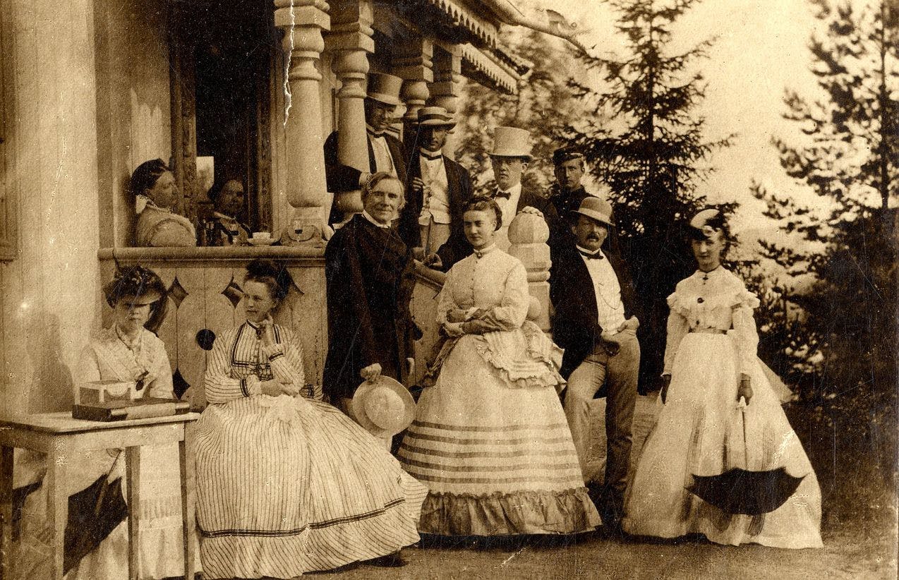 Photo of Ole Bull and his wife Sara Thorp together with friends, all standing outside on the front porch of a house dressed in beautiful clothes. 