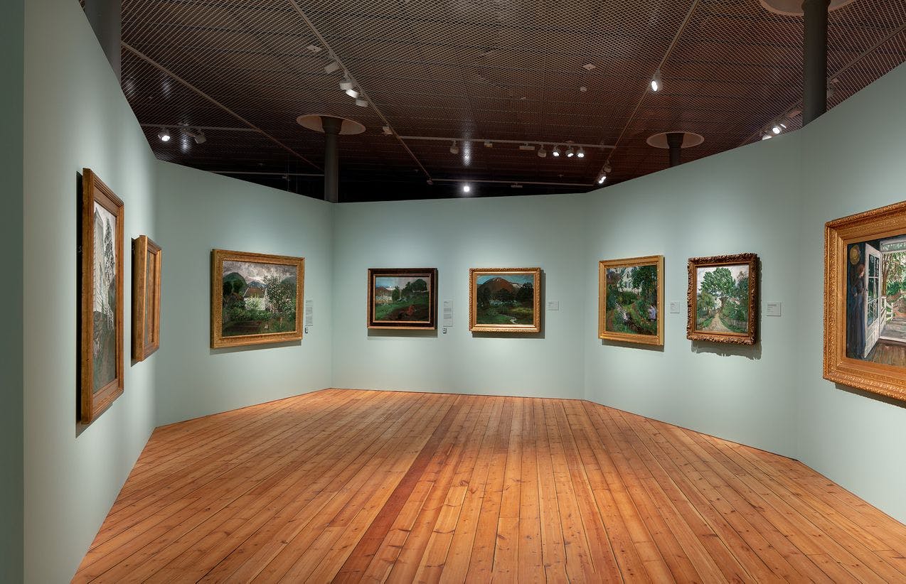 Fotografi fra Astrup-utstillingen som viser en rekke med malerier utstilt på vegg i et buet rom.