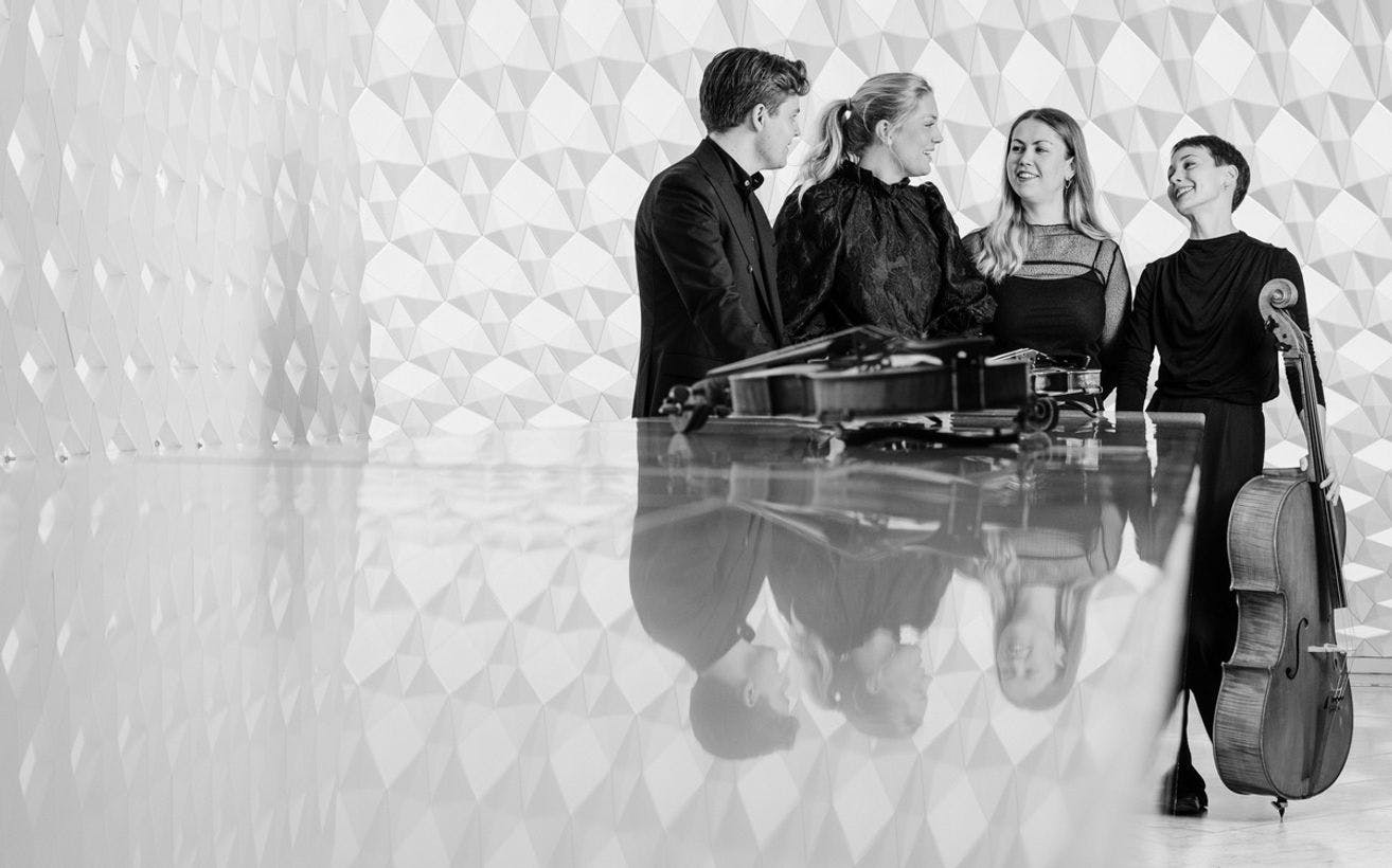 Et sort-hvitt foto av fire musikere. En av dem holder en cello. De ser på hverandre og smiler.