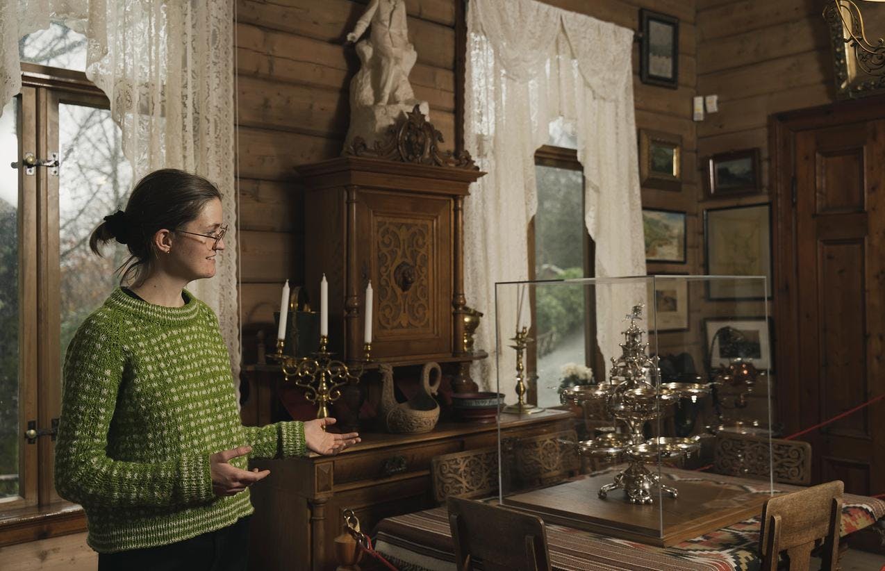 En omviser i Griegs villa peker mot en stor sølvoppsats som er plassert på et bord. Møbler med rike utskjæringer i bakgrunnen.