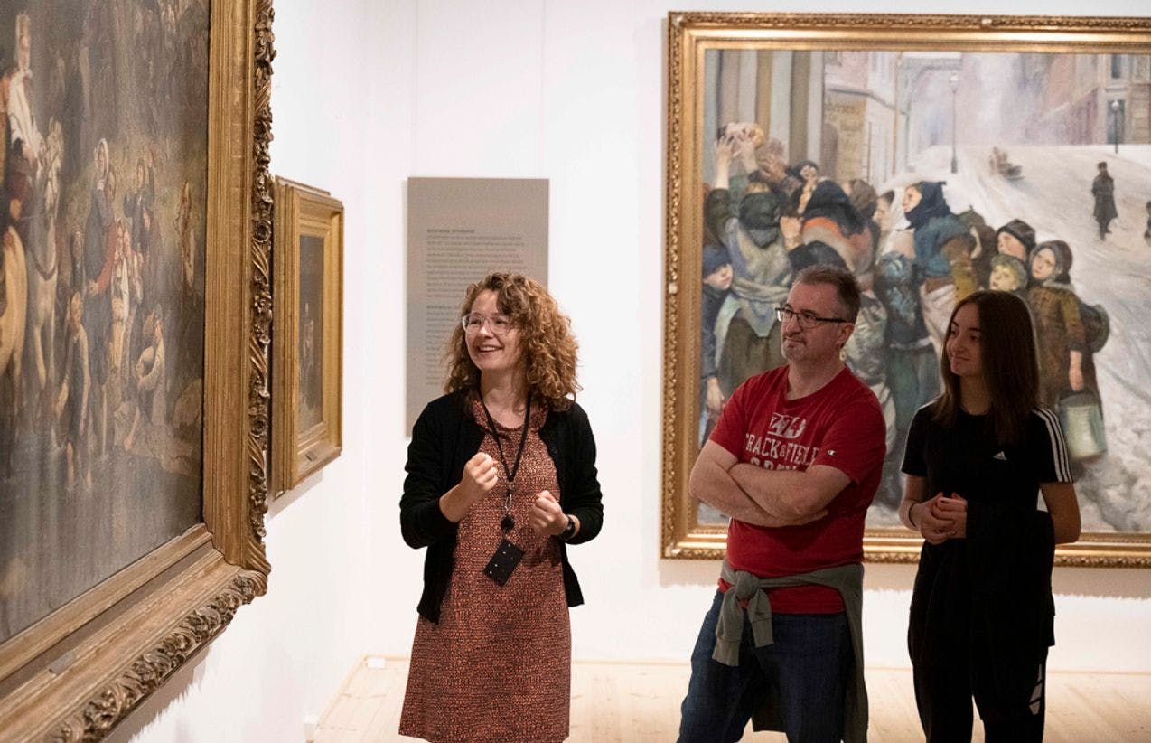A guide is standing in front of a large painting, talking. Behind her are two people listening.