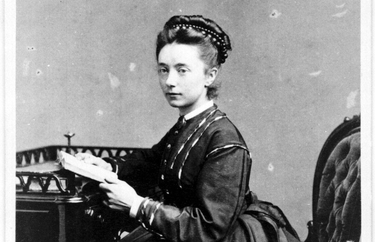 Fotografi av Nina Grieg som ung, sittende ved et skrivebord i lang kjole. 