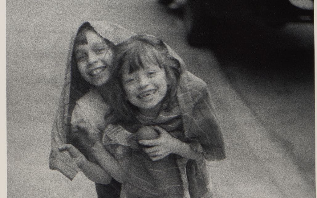 Photography by Diane Arbus depicting two happy children, laughing, and hiding together under a large jacket while walking towards the photographer.