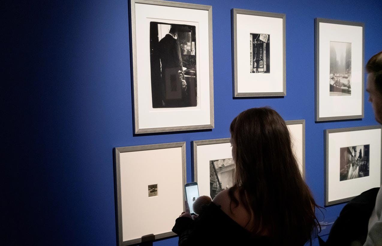 En person i et utstillingsrom med blå vegger, og sort-hvite fotografier. Hun tar bilde av fotografiene.