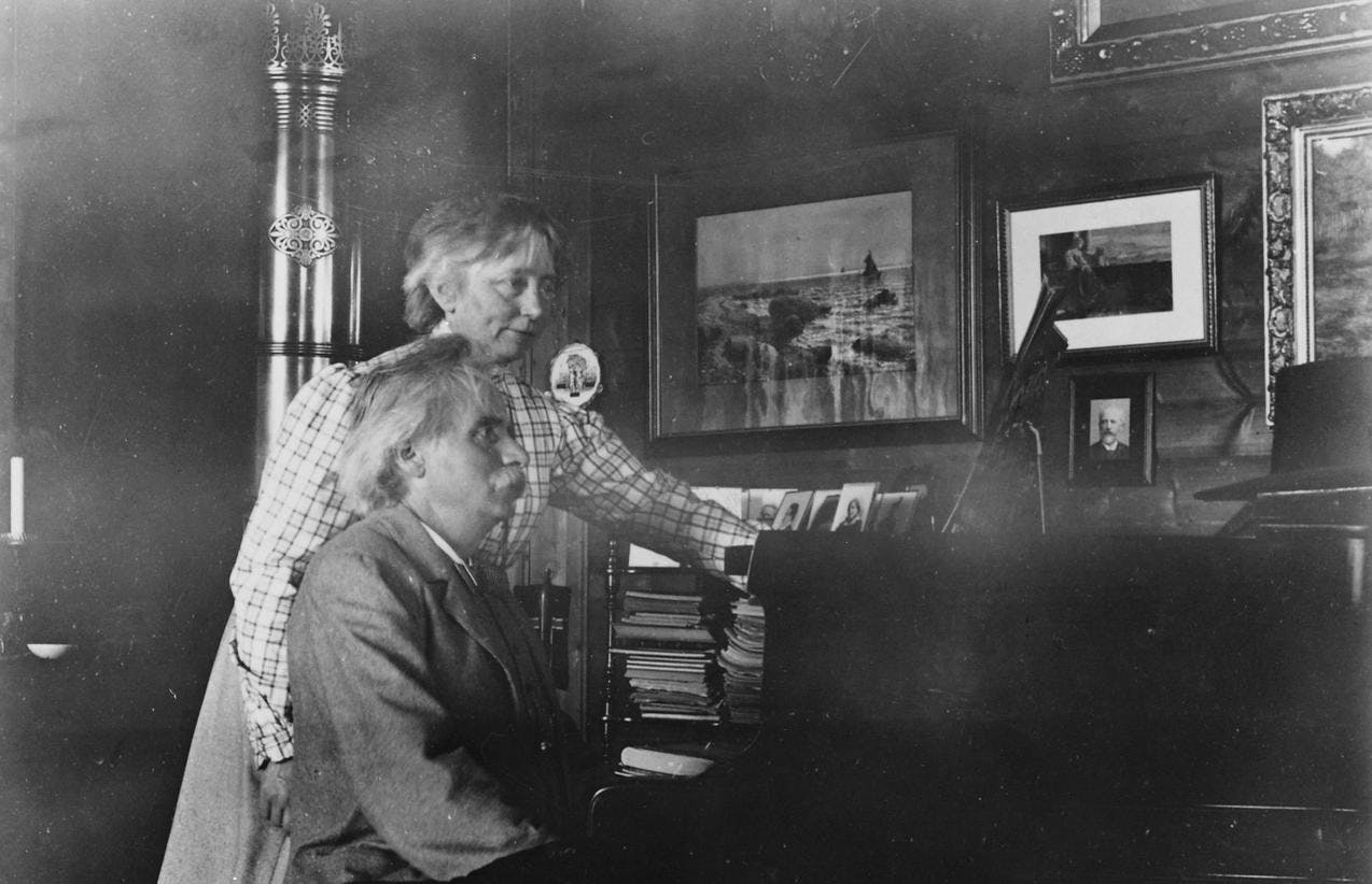 Et gammelt fotografi av Edvard Grieg som spiller pianoet med konen Nina stående bak ham.