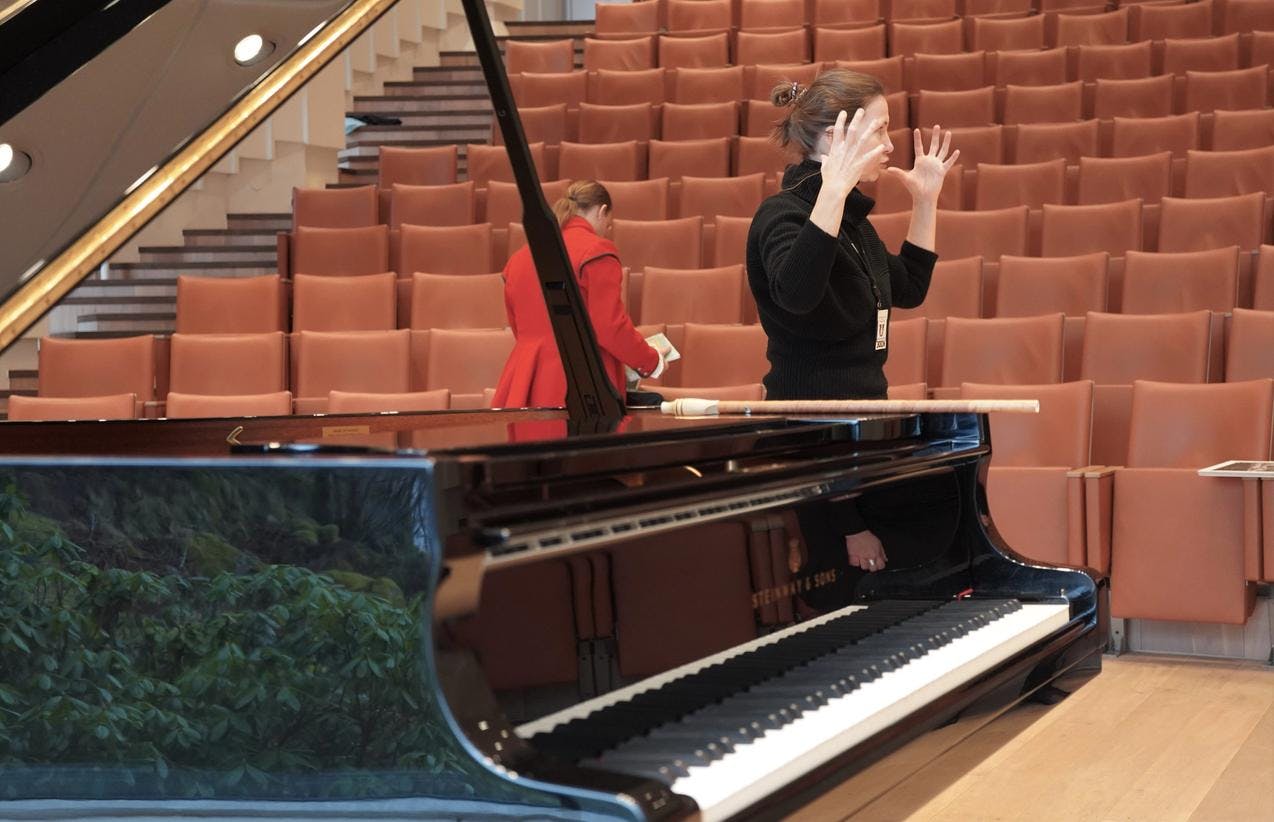 En kvinne gestikulerer med hendene, mens hun står i konsertsalen på Troldhaugen. I forgrunnen ser vi flygelet.