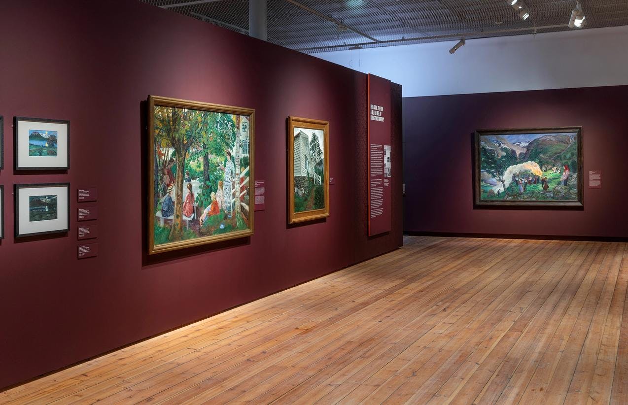 An exhibition room with dark red walls, featuring paintings by Nikolai Astrup.