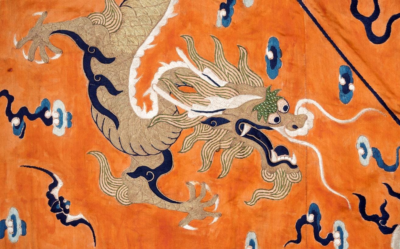 En detalj fra en kinesisk tekstil, som viser et broderi av en drage, mot en gul-oransje bakgrunn.