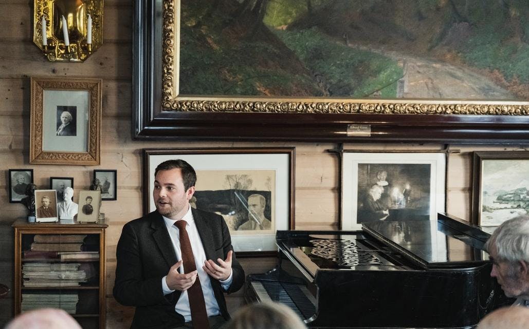 Foto som viser direktør ved komponisthjemmene, Christian Grøvlen, sittende ved pianoet i Griegs villa. Han forteller noe til publikum. 