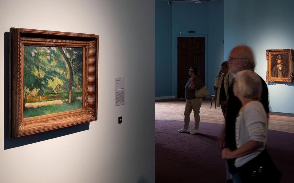 Fotografi fra Cézanne-utstillingen hvor vi ser en eldre kvinne og mann som ser på et maleri av et skogslandskap. I bakgrunnen ses to andre mennesker og noen bilder på vegg.