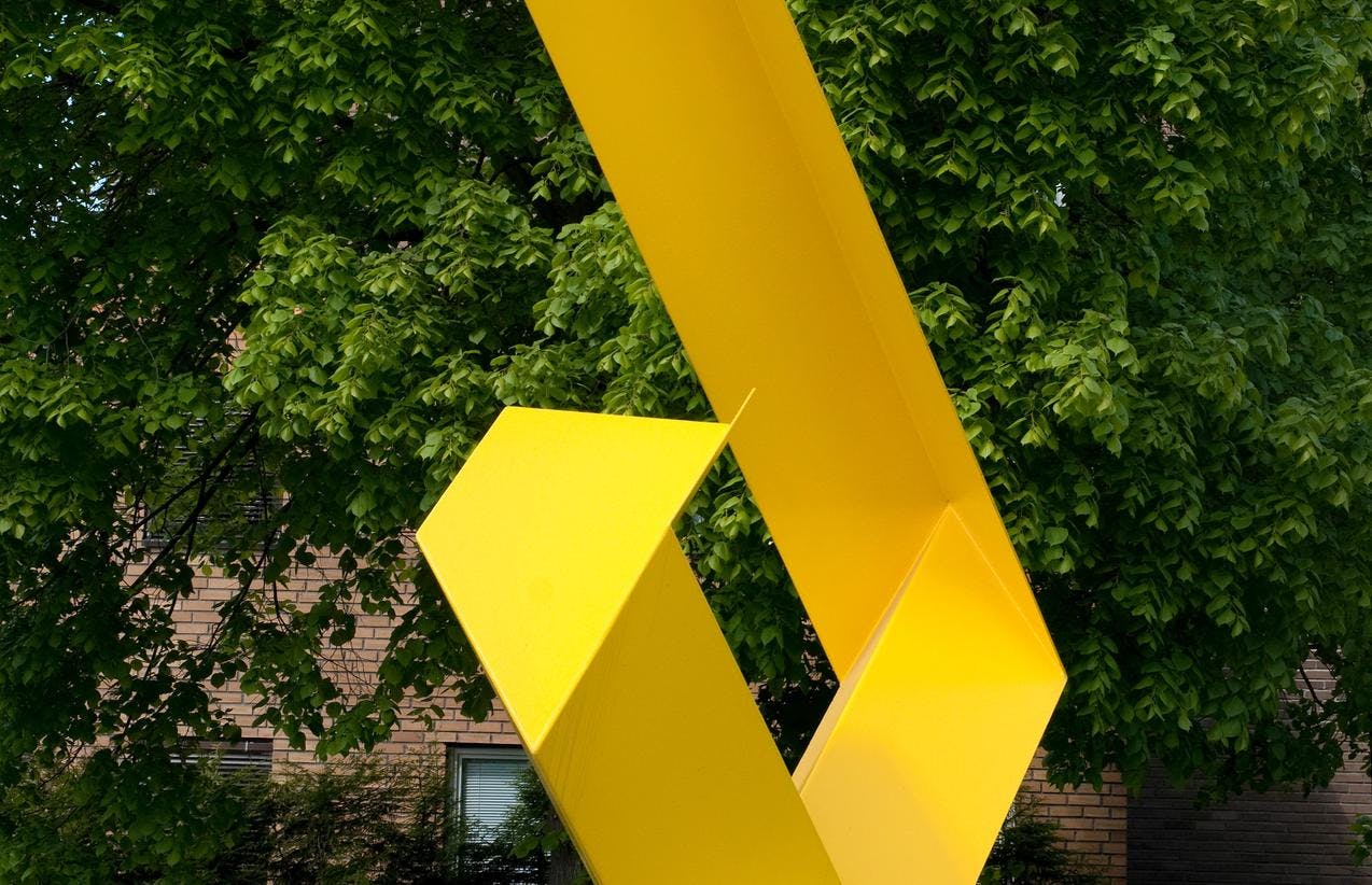 En gul og geometrisk skulptur fotografert i en hage.