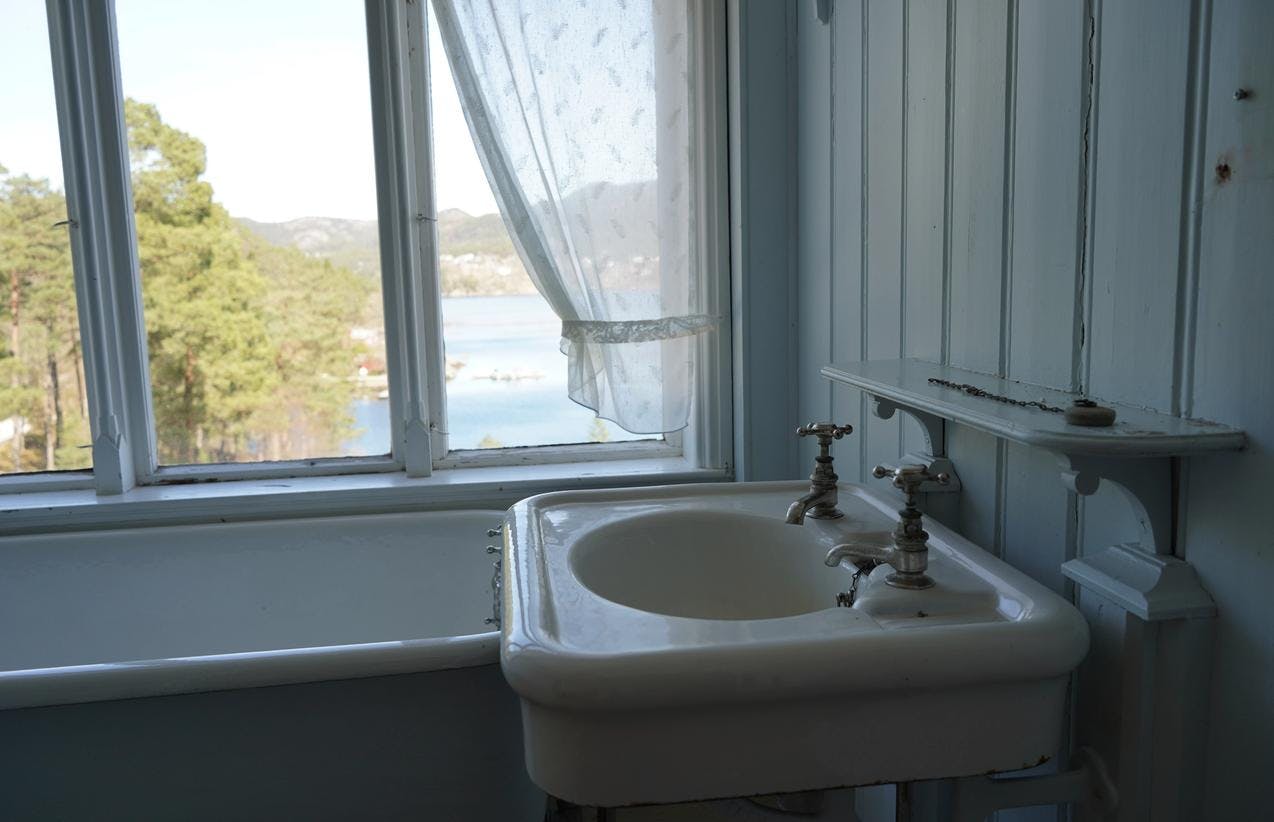 En hvit vaskeservant og badekar, foran et vindu, med utsikt mot trær og sjø