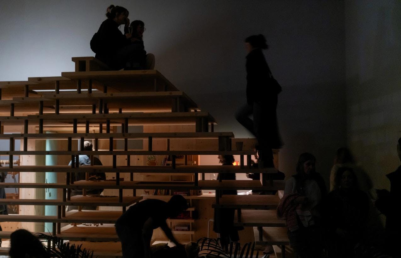 Et foto fra utstillingen Nabo som viser en stor hyllestruktur i tre, med mennesker som sitter på den. 