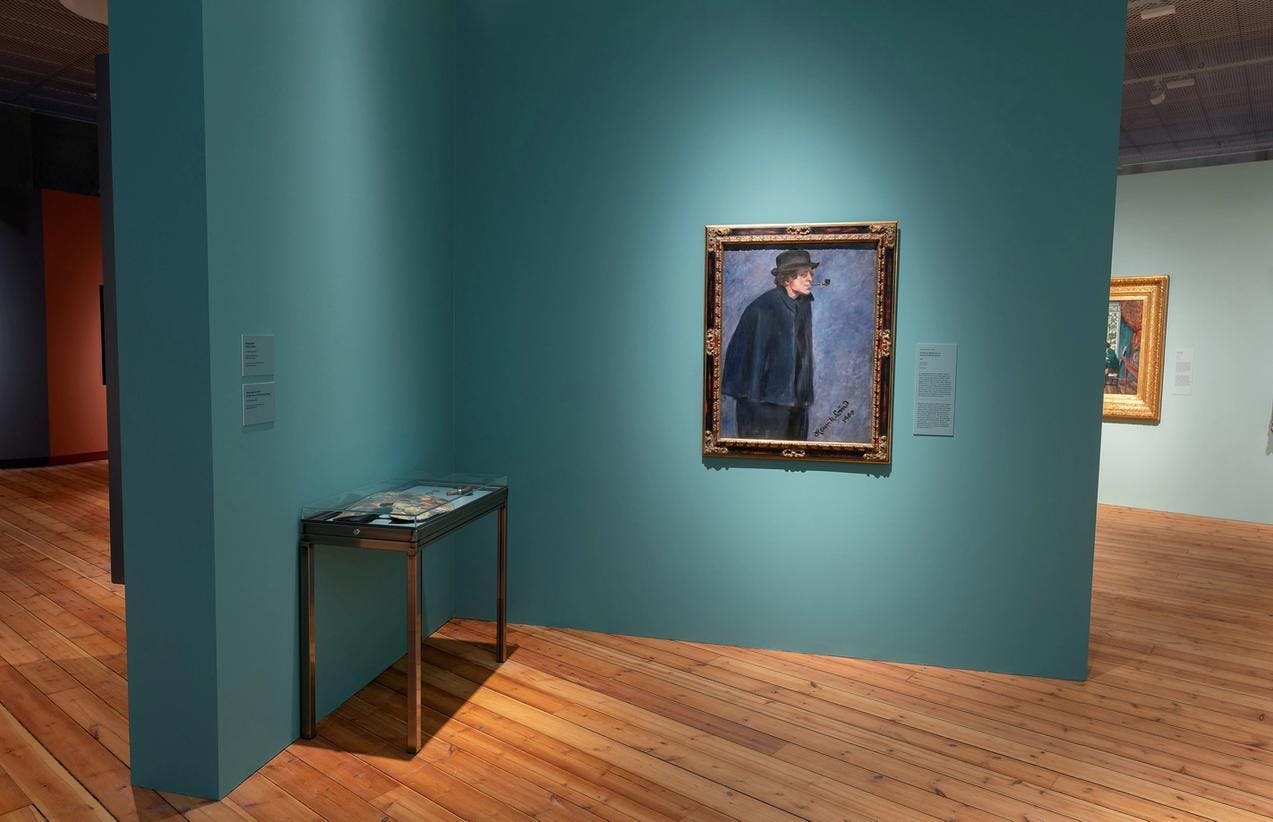 Fotografi fra Astrup-utstillingen som viser en krok hvor et portrettmaleri av kunstneren er hengt på vegg og et bord med gjenstander står ved siden. 