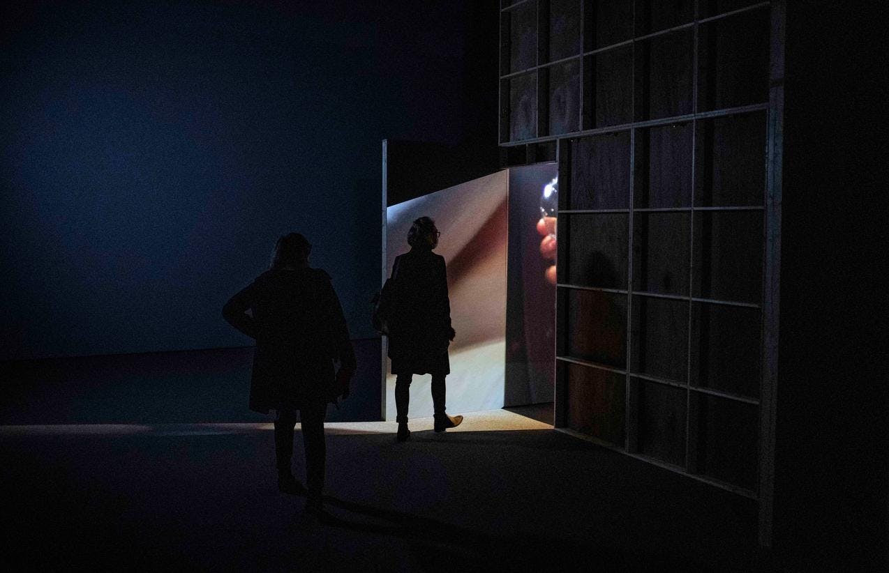 Bilde fra installasjonen DADDA av Paul McCarthy. To mennesker går mot en dør hvor det siver lys ut, rommet utenfor er mørklagt.