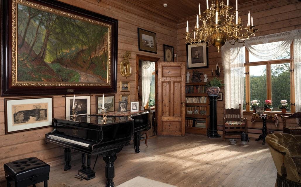 Et foto av Nina og Edvard Griegs stue på Troldhaugen. Interiøret er preget av laftede trevegger, tregulv og møbler i plysj og jordtoner. En lysekrone i messing henger i taket og vi ser Griegs Steinway-flygel som står midt i rommet. På veggen henger en rekke ulike fotografier og malerier fra Griegs tid.