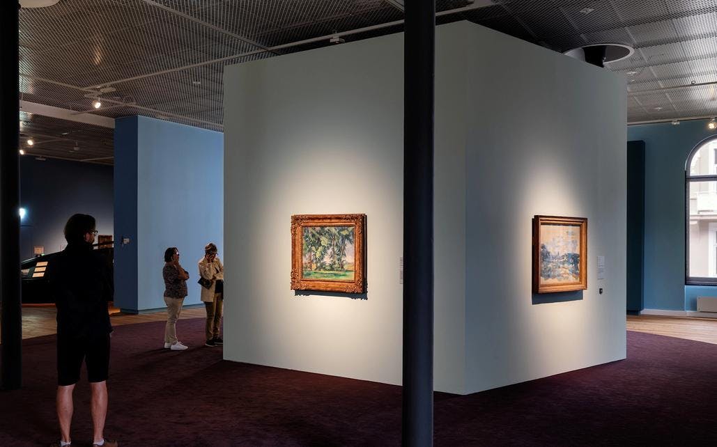 Fotografi fra Cézanne-utstillingen hvor vi ser et overblikk fra lokalet som er stort og åpent med store buede vinduer. I midten er det en veggboks med fire sider, hvor det henger et maleri på hver vegg. Vi ser en gruppe mennesker som går gjennom lokalet. En mann sitter på en benk i bakgrunnen.