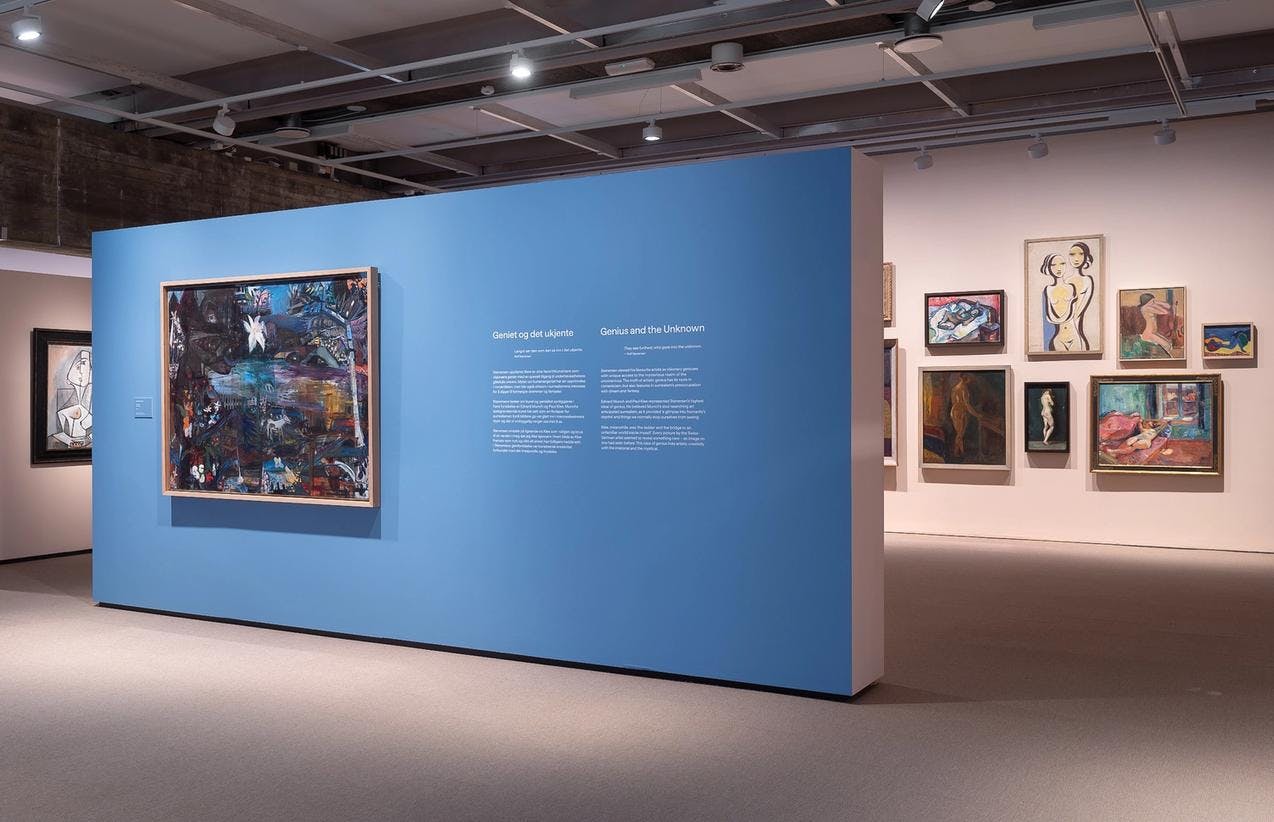 Et utstillingsrom hvor vi ser en blåmalt vegg midt i rommet, omgitt av lysrosa vegger hvor det henger en rekke malerier