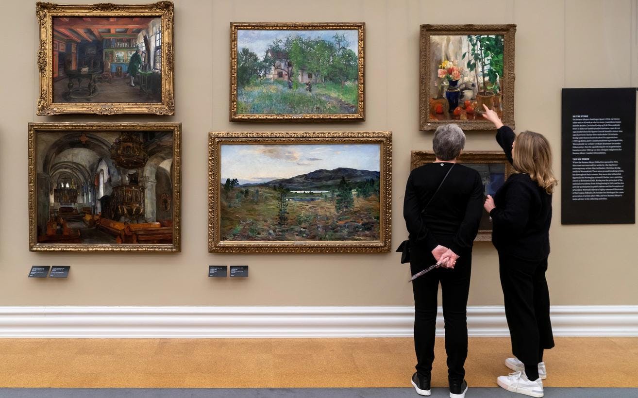 To kvinner står i en utstilling og ser på malerier i gullramme. De ses bakfra. 