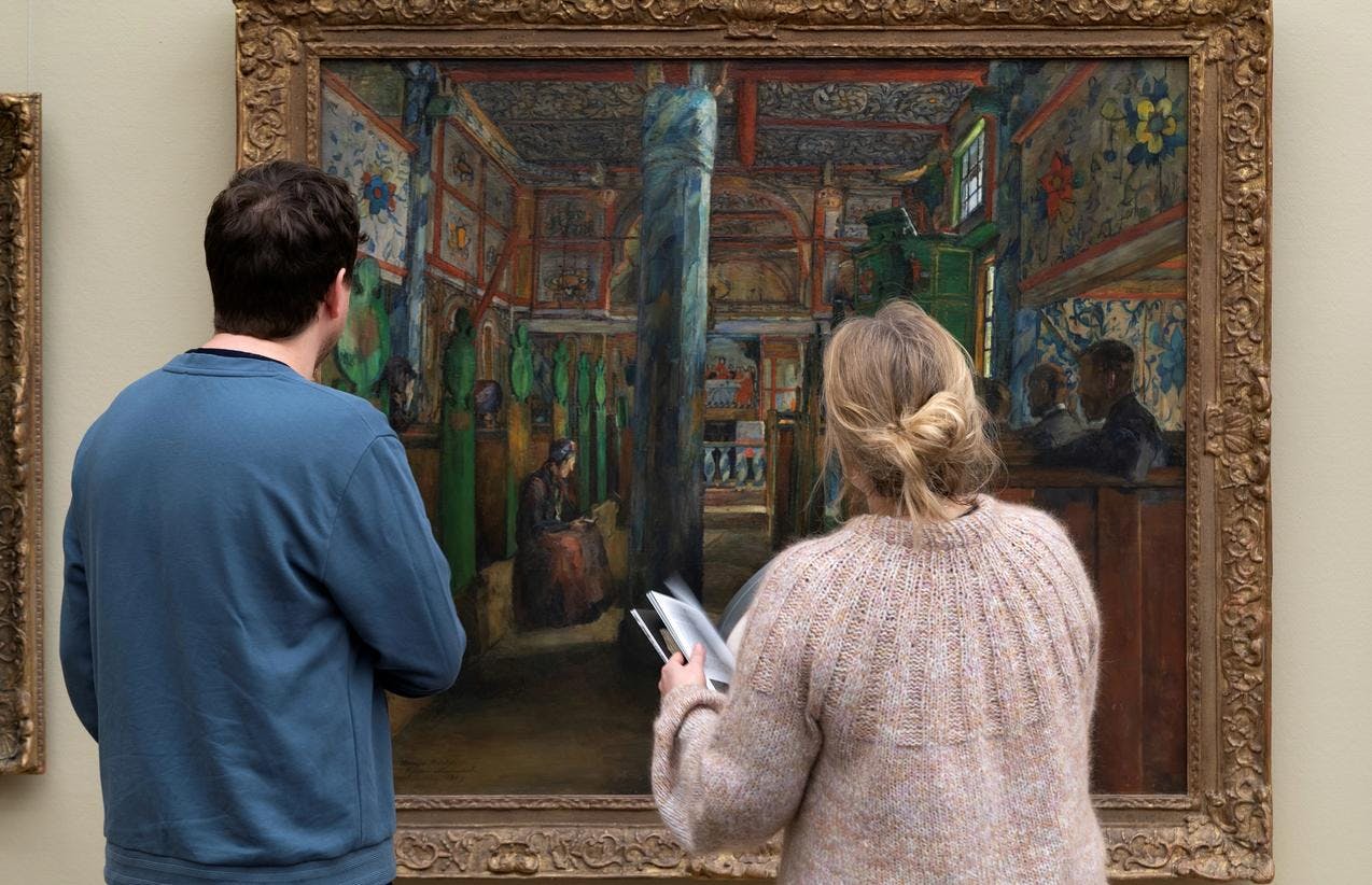 To mennesker står med ryggen mot oss, og titter på et stort maleri i grønntoner malt av Harriet Backer. 