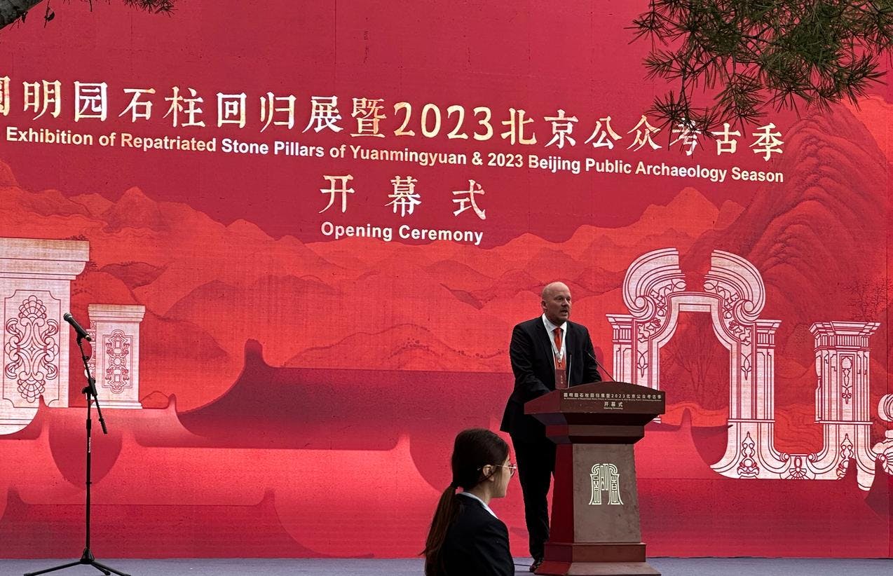 En mann holder tale på et podium foran en rød bakgrunn hvor det står "åpningsseremoni" på kinesisk.