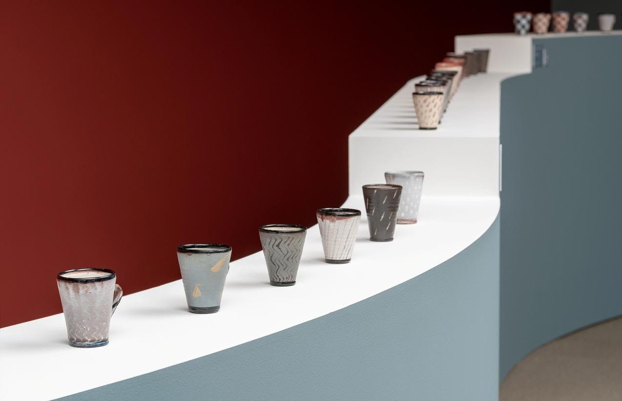 Fotografi fra utstillingen Materielle påstander av Nina Malterud. Vi ser en rad med kopper i keramikk som står på en buktende søyle. 
